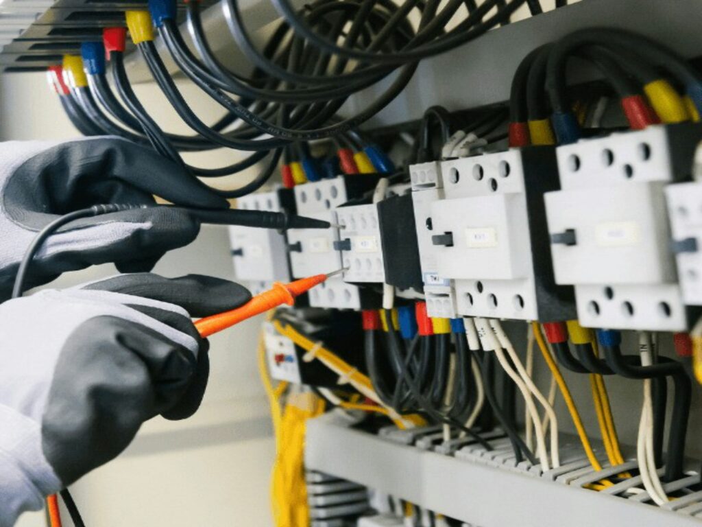 Electrician wearing protective gloves testing electrical circuits inside a control panel with multiple wires and breakers